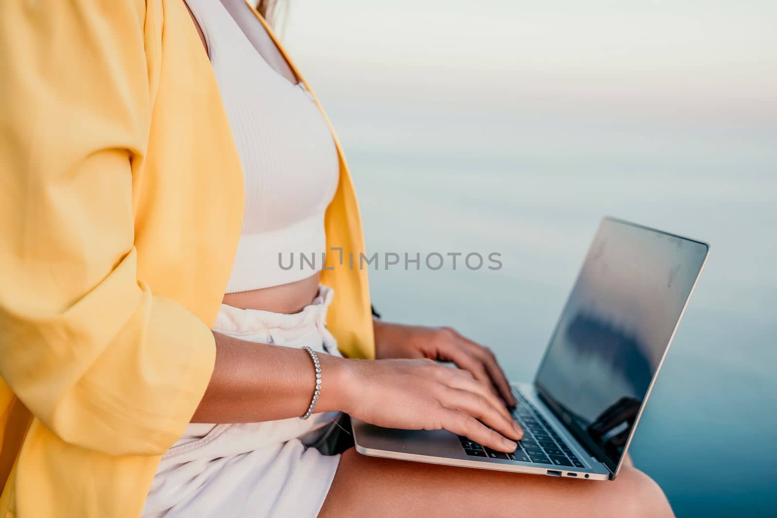 Digital nomad, Business woman working on laptop by the sea. Pretty lady typing on computer by the sea at sunset, makes a business transaction online from a distance. Freelance, remote work on vacation by panophotograph