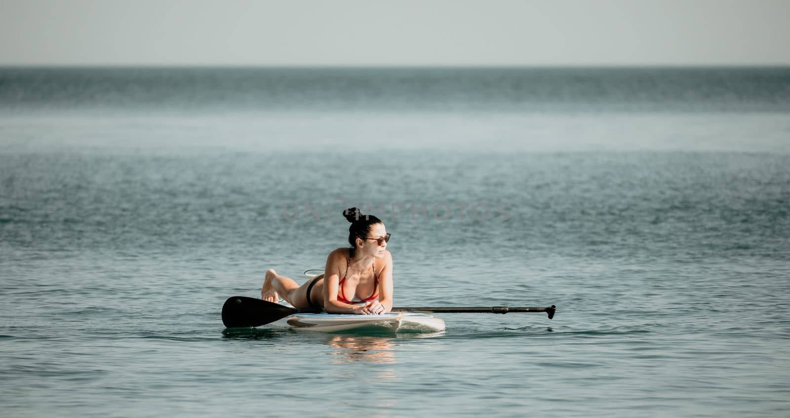 Sea woman sup. Silhouette of happy middle aged woman in rainbow bikini, surfing on SUP board, confident paddling through water surface. Idyllic sunset. Active lifestyle at sea or river