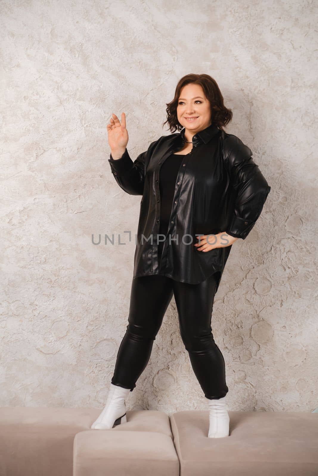 An adult woman in black leather clothes poses against a gray studio wall.