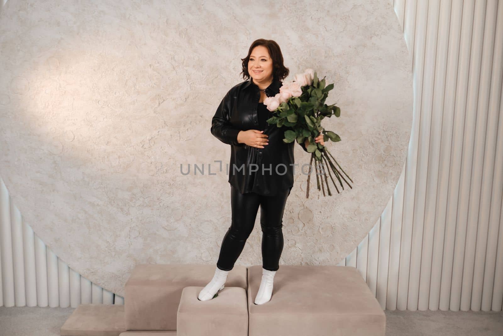 a stylish adult woman in black leather clothes stands with a bouquet of pink roses in the interior.