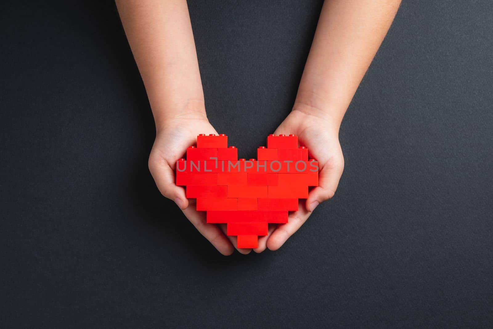 Hands of little girl child holding heart made of plastic bricks on dark gray background