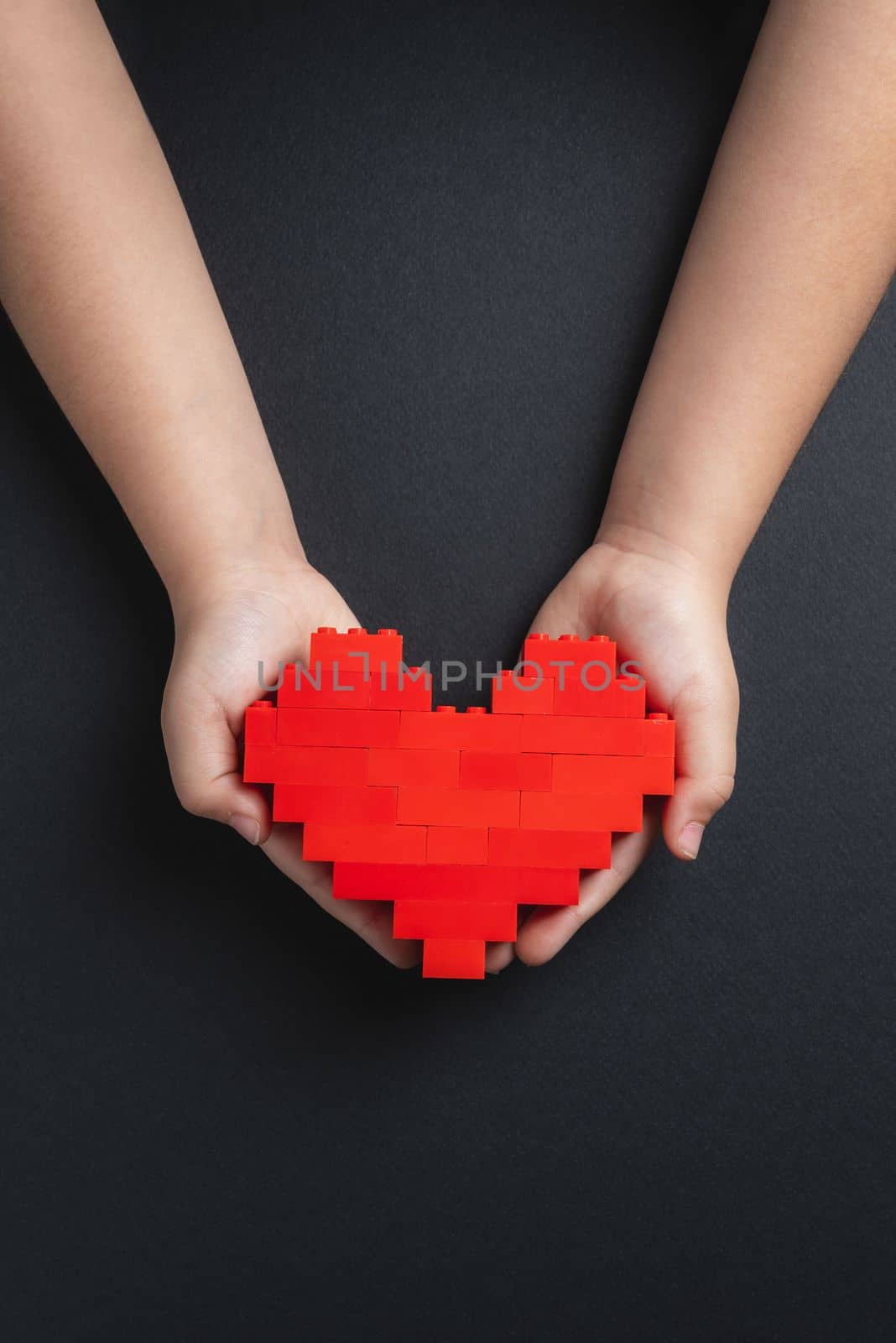 Hands of little girl child holding heart made of plastic bricks on dark gray background