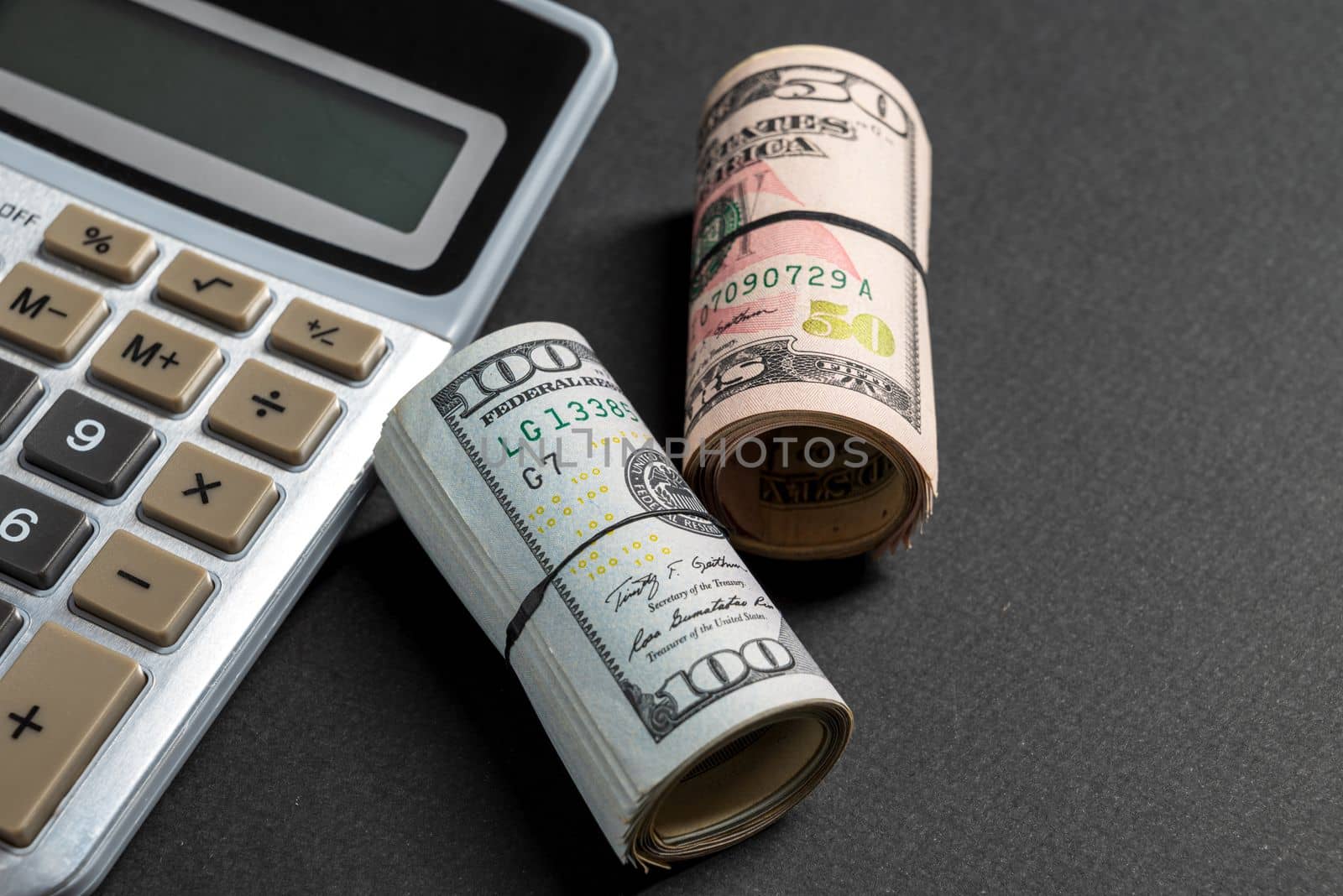 Rolls of 50 and 100 us dollar standing near calculator on dark background by Sonat