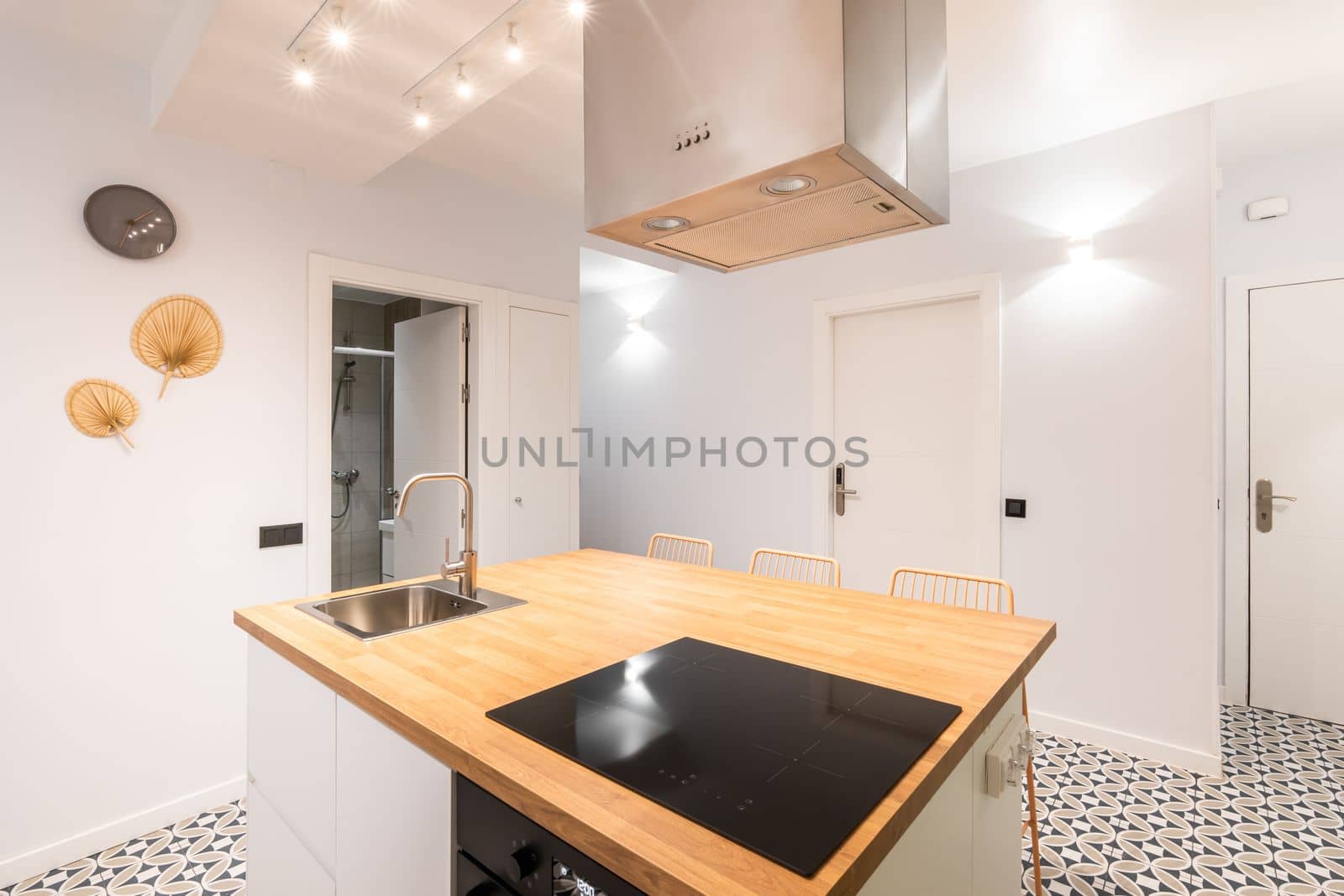 An island table in spacious studio kitchen in modern apartment with trendy author's design. On wooden surface there is an induction electric stove above it with an exhaust system. Mosaic tile floor. by apavlin