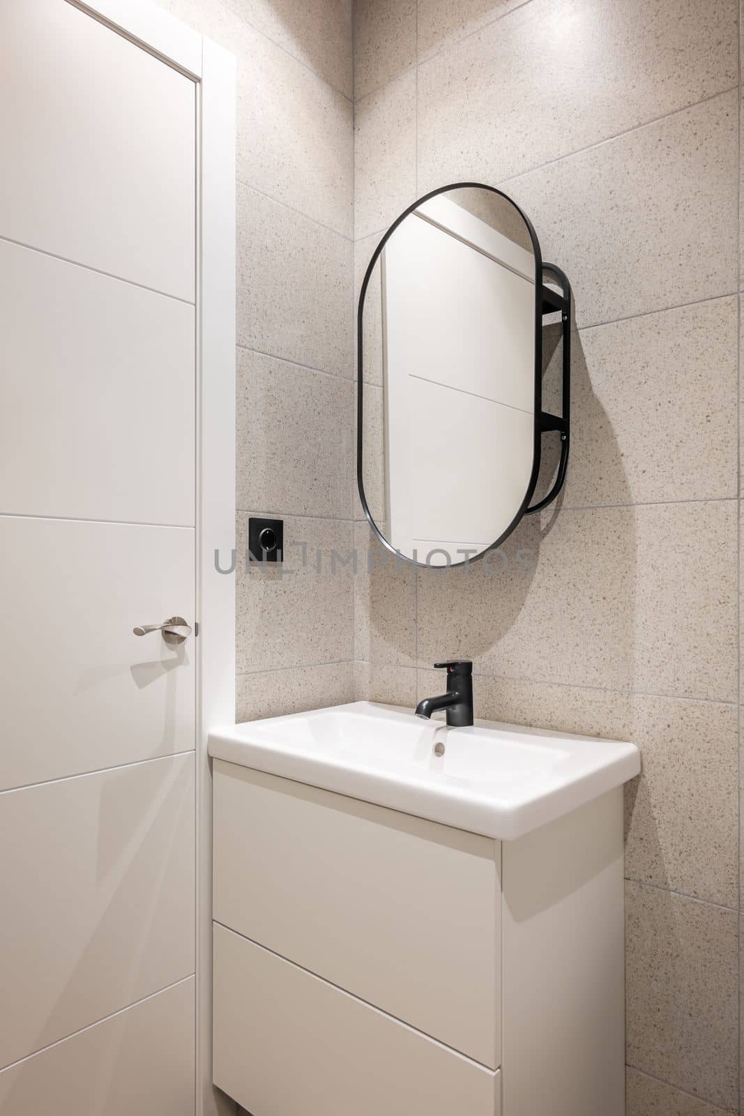 Corner of a bathroom with a white sink on a vanity with two large drawers for bathroom amenities. Above the sink is an oval designer mirror. Decorative elements made of black metal. by apavlin