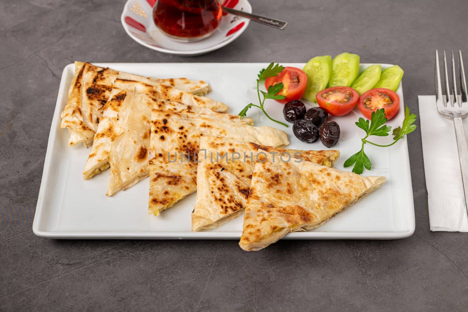 Traditional Turkish flatbread or Gozleme with tomatoes, olives and tea on dark stone table by Sonat