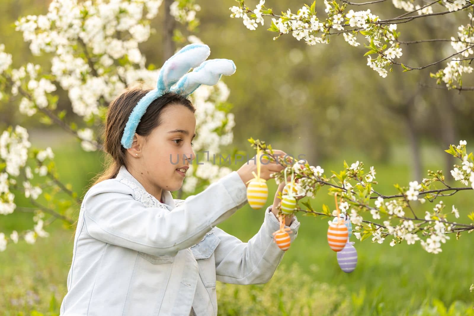 little girl wearing bunny ears with colorful Easter eggs outdoors on spring day.