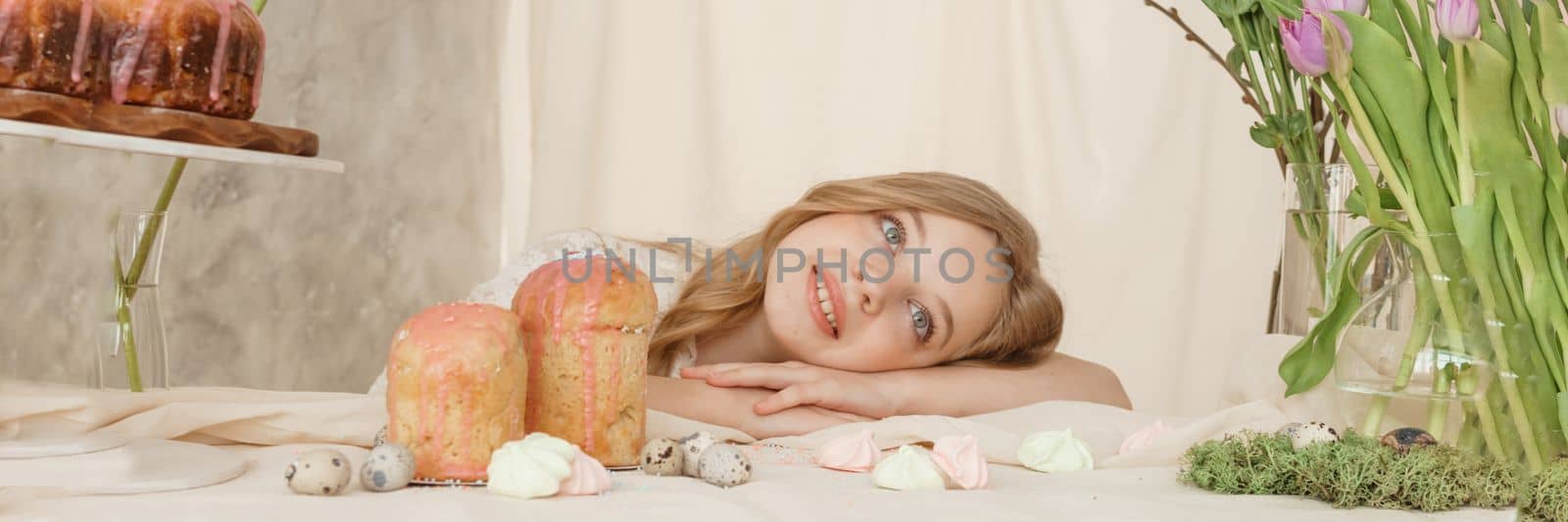 A girl with long hair in a light dress is sitting at the Easter table with cakes, spring flowers and quail eggs. Happy Easter celebration