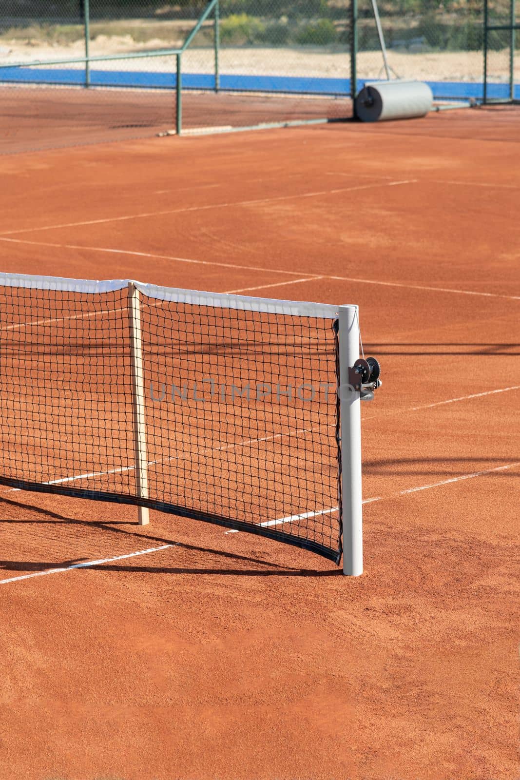 Baseline and net of an empty clay tennis court on a sunny day by Sonat