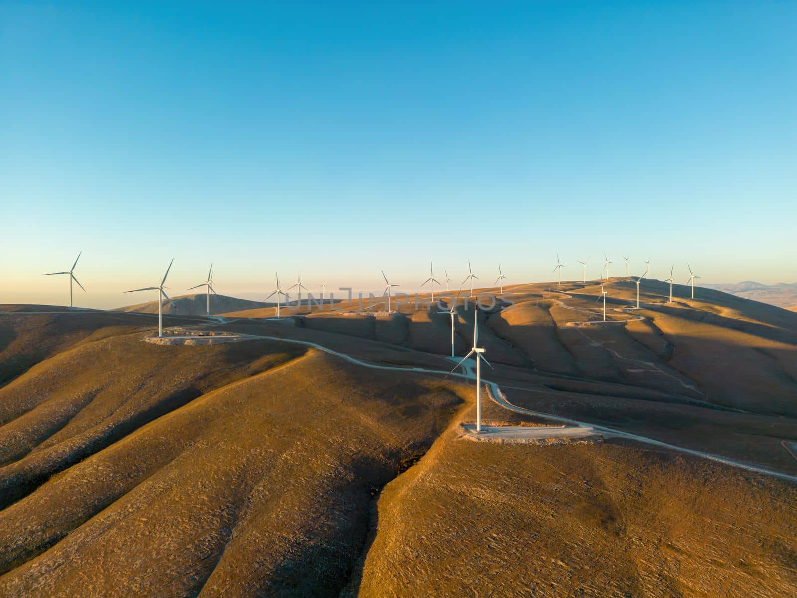 Aerial view of multiple wind turbines standing on a hill and generating electricity at sunrise by Sonat