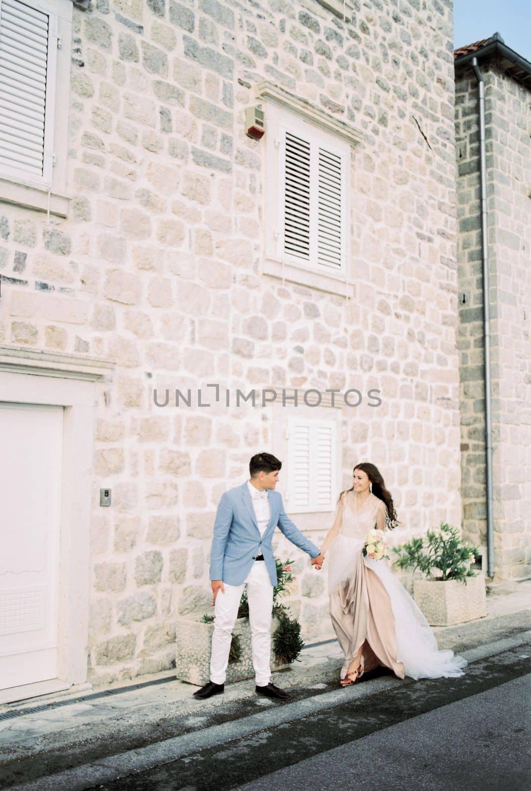 Bride and groom walk hand in hand past an old stone building. High quality photo