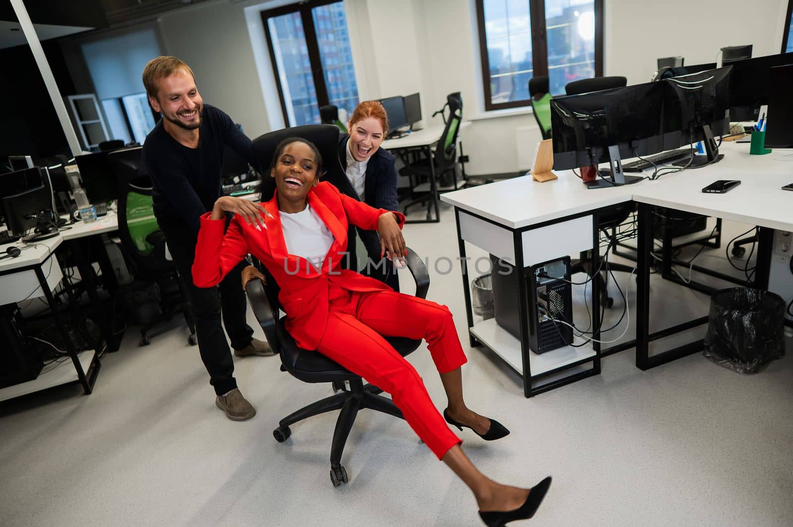 Caucasian red-haired woman, bearded caucasian man rolled African American young woman on office chair. Colleagues have fun at work