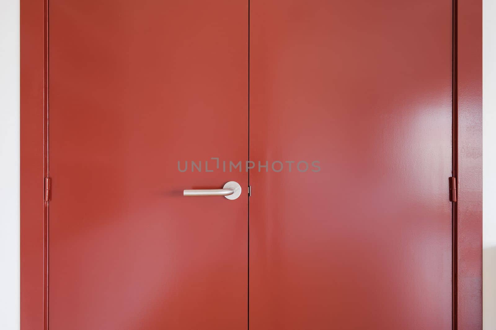 Closeup of a wide front double-leaf wooden door covered with red paint. Door handle made of durable metal. The door is held on door hinges. Sunlight sets off the depth of color