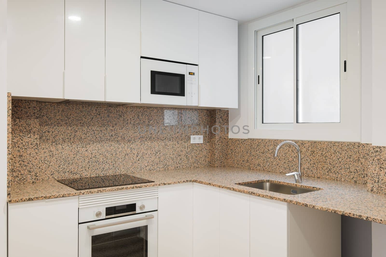 Modern kitchen is well lit by daylight from a sliding sash window. Beige granite walls with black spots. Kitchen furniture made of wood covered with white glossy paint. Built-in modern technology. by apavlin