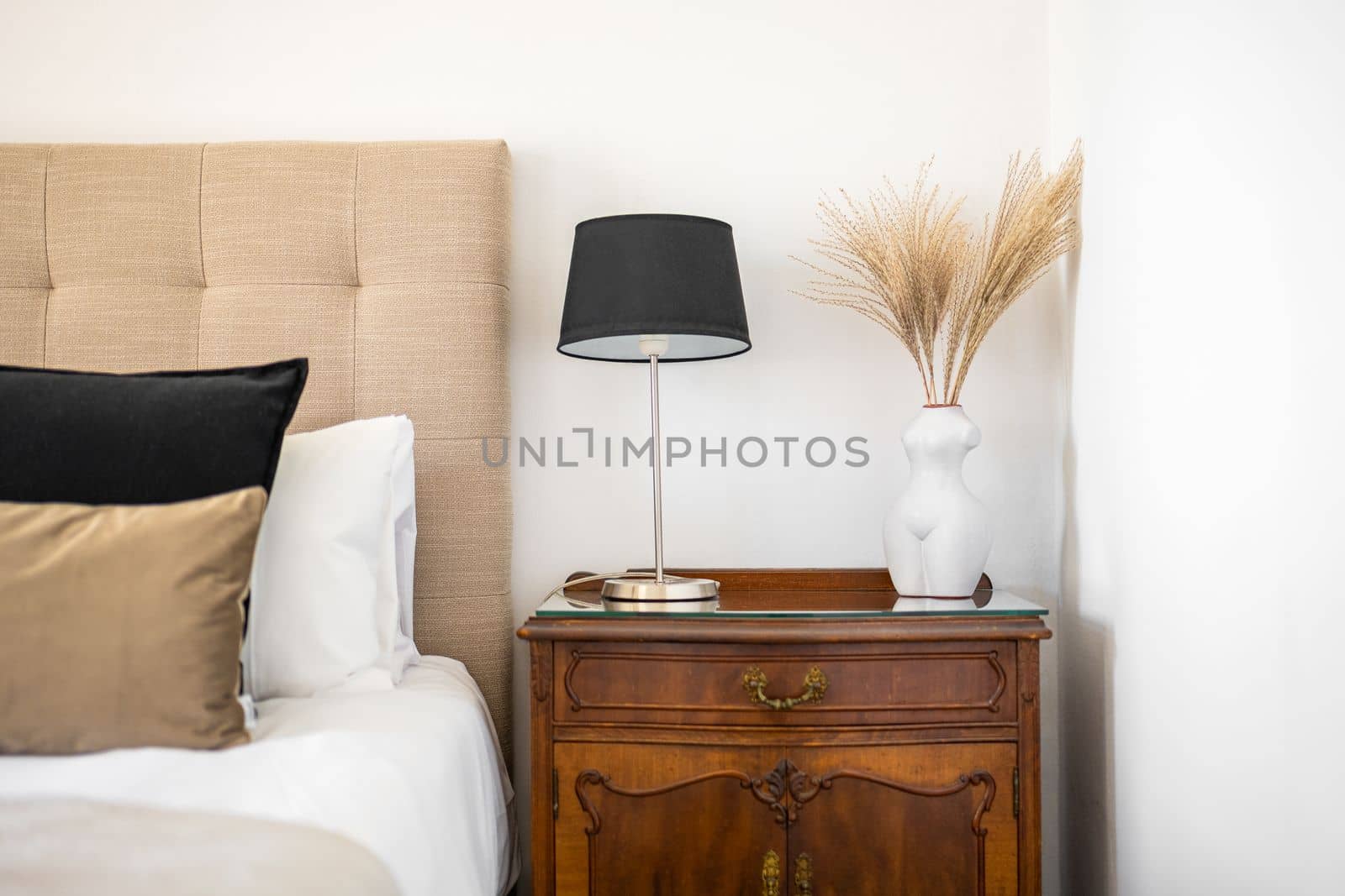 Chic bedroom interior with baroque bedside table with complex ornaments and monograms. On bedside table is night lamp with black shade and vase with decorative dried flowers. Bed with velor pillows