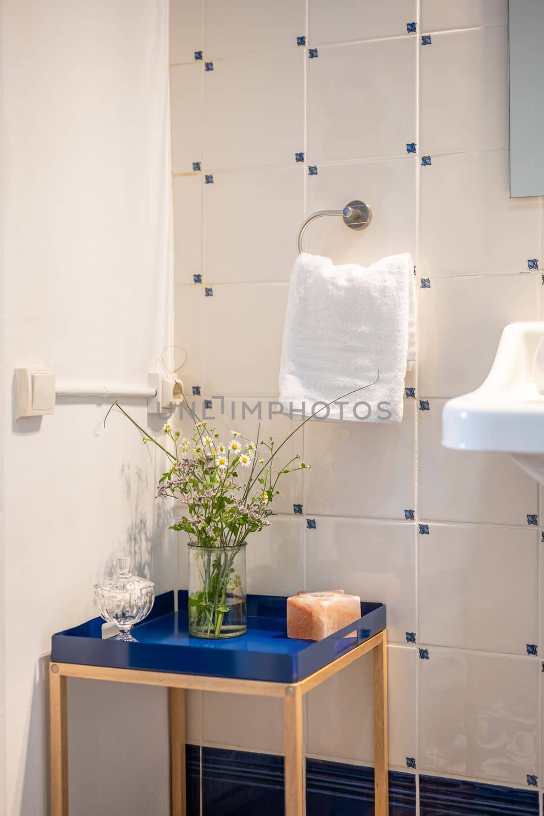 Corner of a bathroom with a decorative low decorative table with a blue base. On the table is a vase with wild flowers. On a tiled wall is a holder with a white terry towel