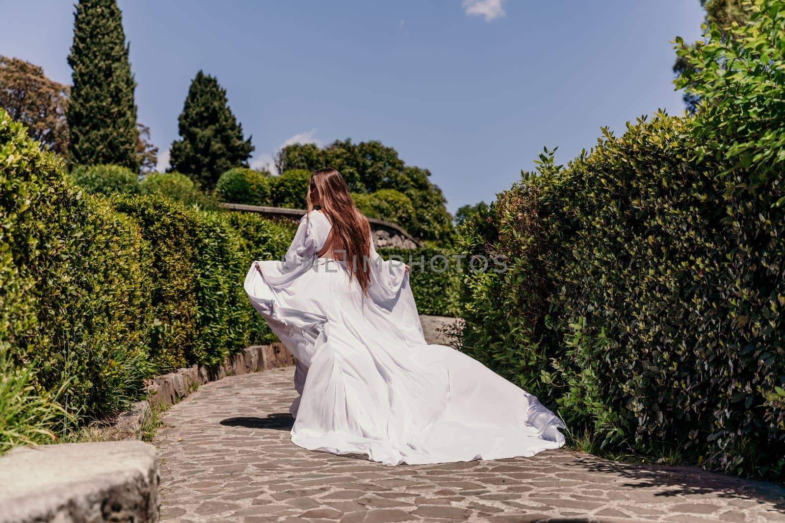 Brunette runs white dress park. A beautiful woman with long brown hair and a long white dress runs along the path along the beautiful bushes in the park, rear view.