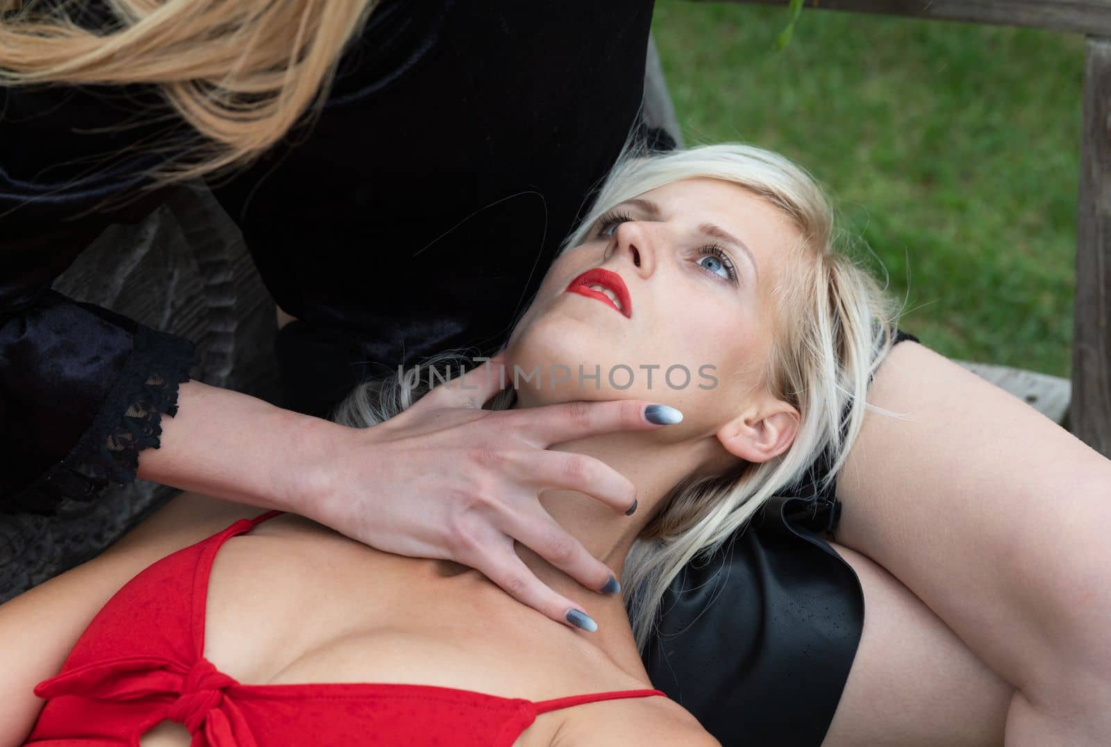 Romantic relationship. Two young women on outdoor wooden swings. A blonde girl in red is lying on the lap of a brunette girl in a black dress who has her hand on the blonde's throat