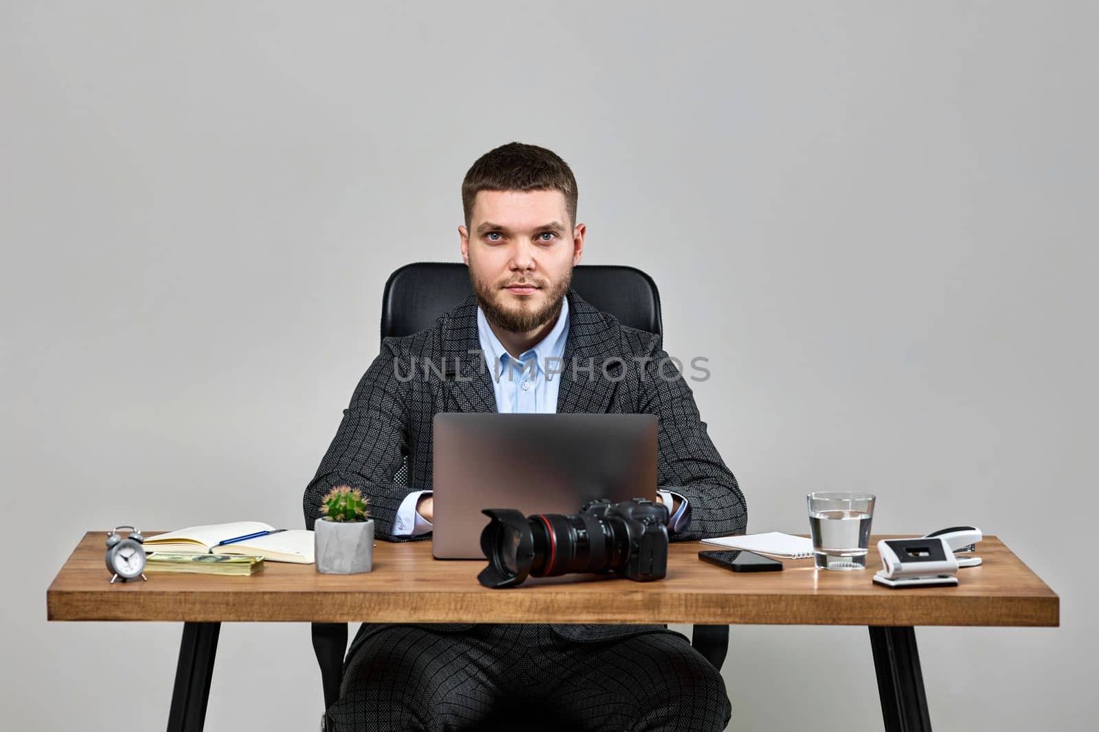 bearded male photographer smiling cheerfully while working on laptop computer at desk. dslr camera on table
