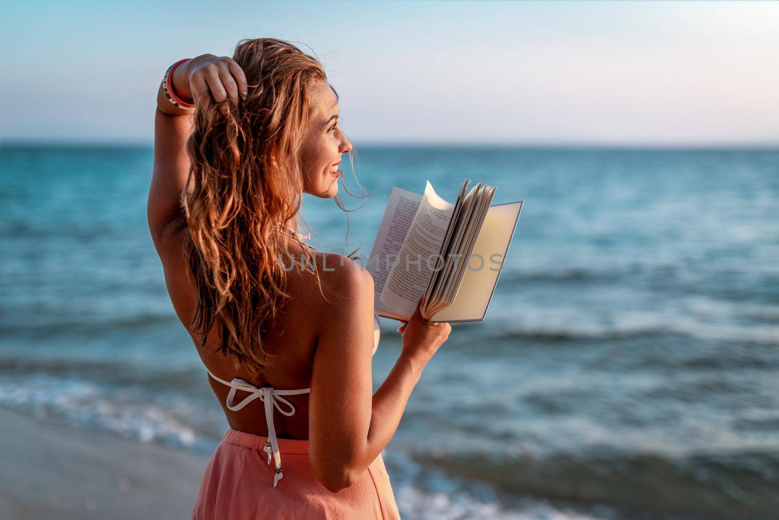 Young woman holding a book on the beach. She is standing next to the water and looking away with smile on her satisfied face.