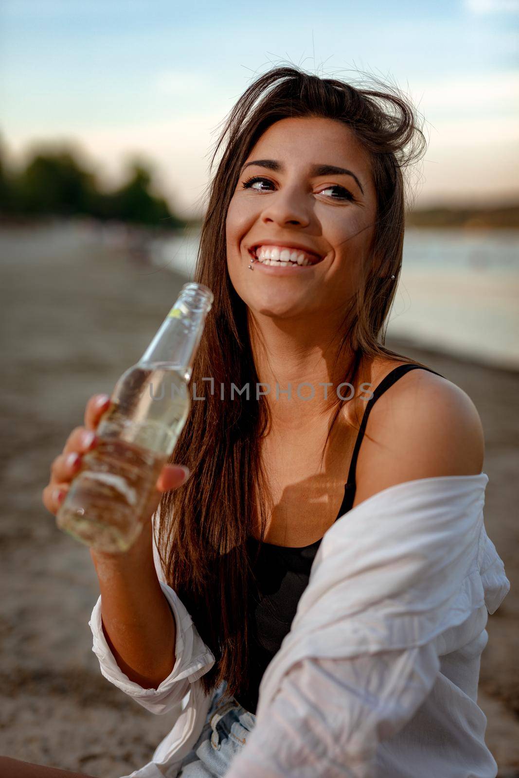 Young woman relaxing at sunset time on the river bank. She is sitting by the river and drinking beer. 