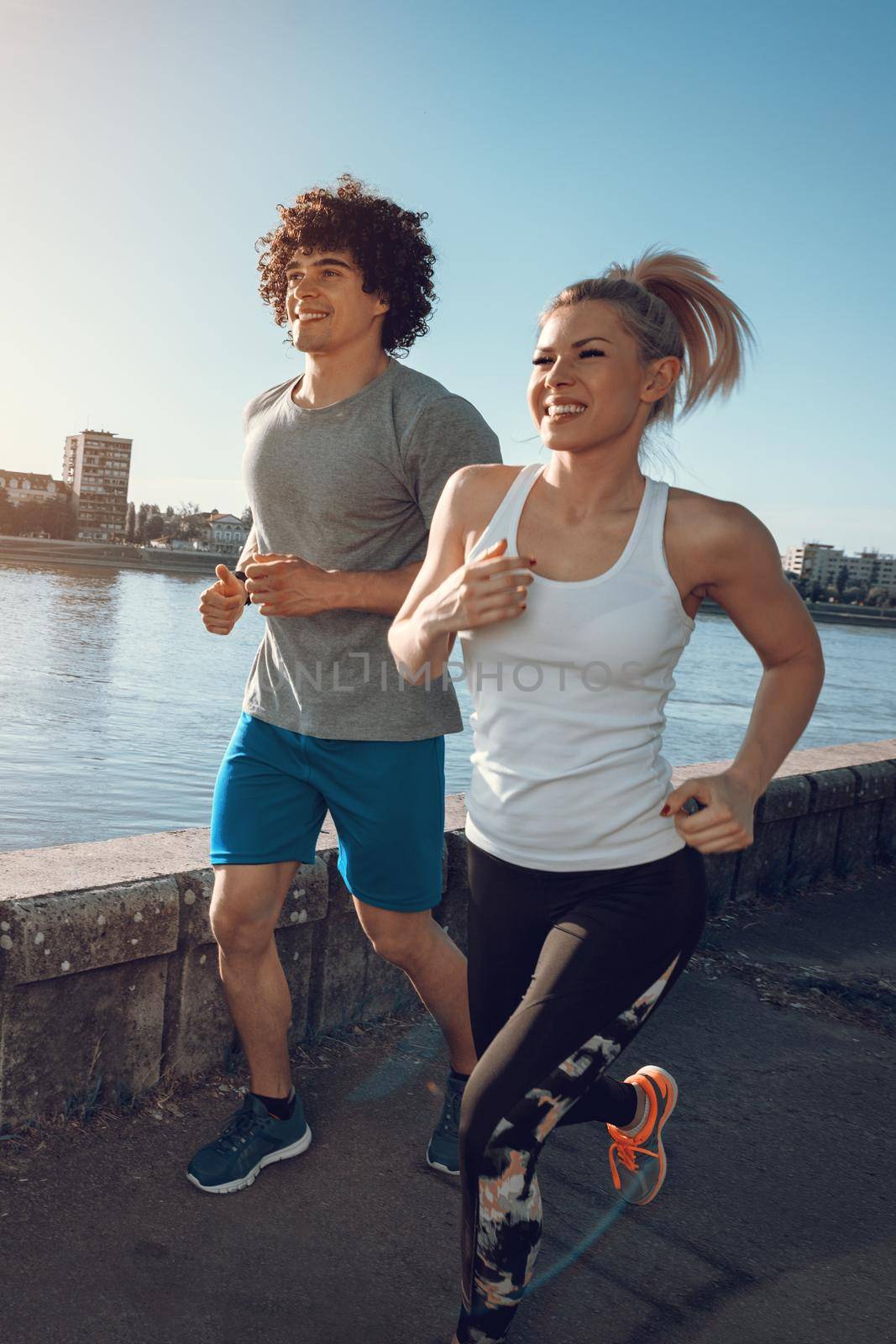 Young happy runners couple training outdoors by the river, working out in nature against blue sky with sunrise light.