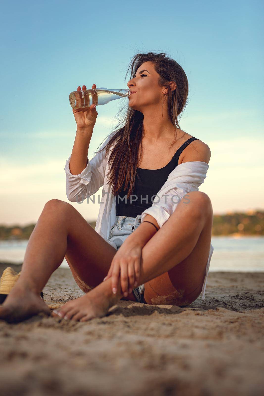 Young woman relaxing at sunset time on the river bank. She is sitting on the river send and drinking beer. 