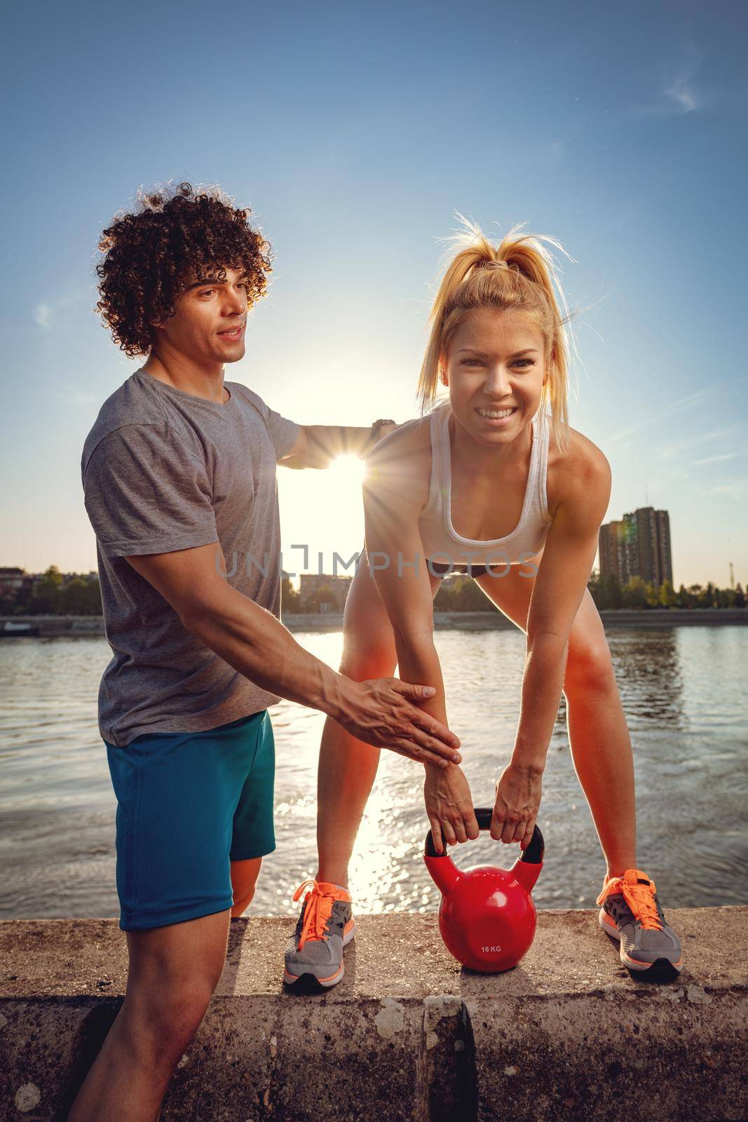 Young fitness couple doing workout with kettlebell on the wall  by the river in a sunset. The woman is crouching and holding kettlebell, and the man support her.