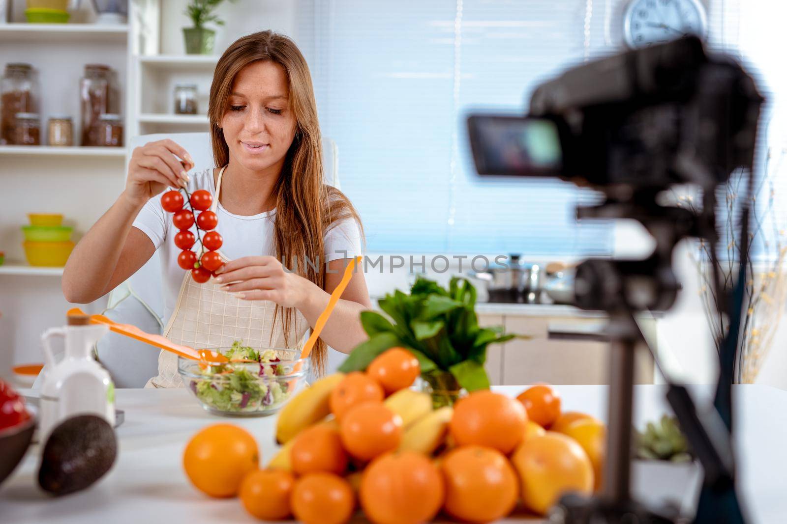 Beautiful young woman filming her blog broadcast about healthy food at home. She is holding cherry tomato in her hand and putting it in a salad.