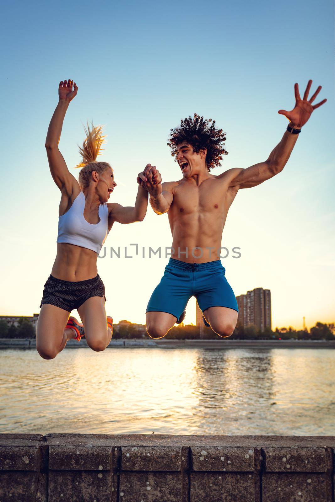 Beautiful young couple smiling and jumping after successful training by the river in the sunset.