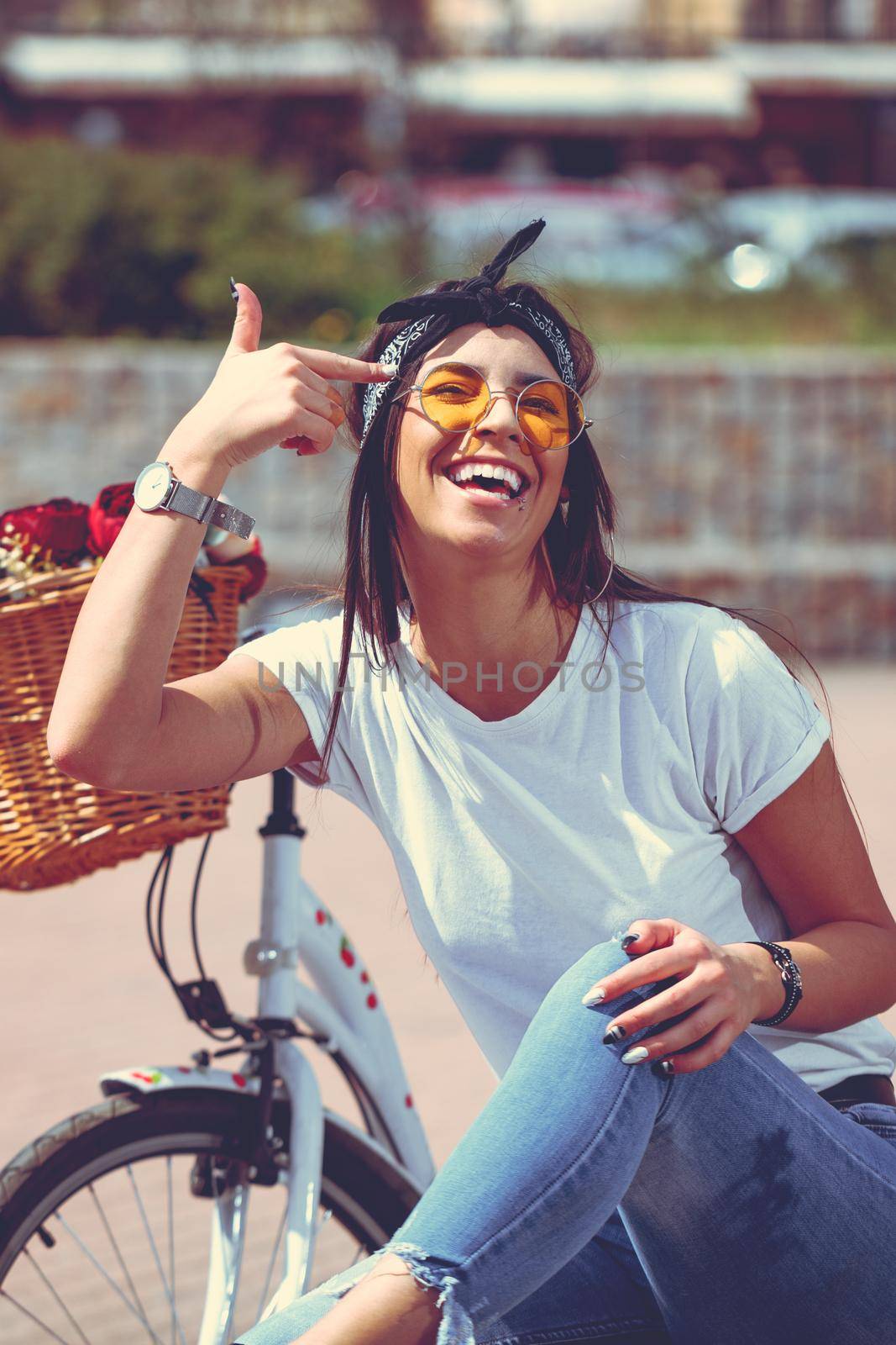 Pretty happy young woman is enjoying and having fun putting a finger on her forehead, in a summer sunny day, beside the bike with flower basket.