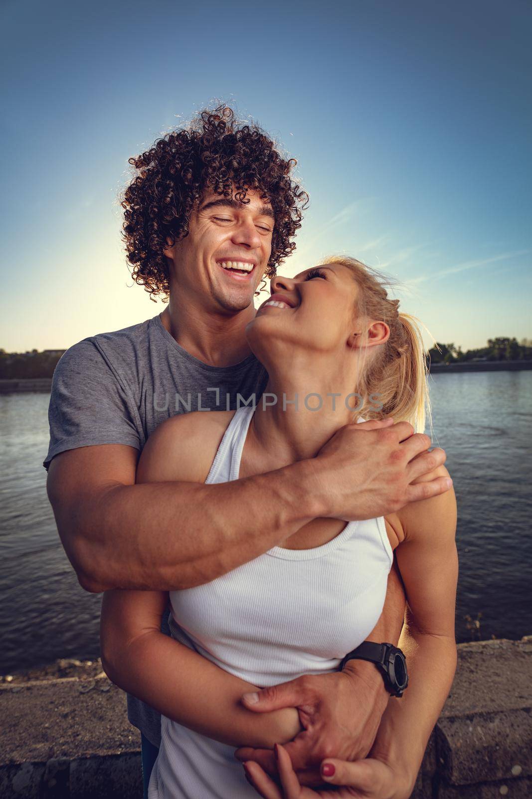 Beautiful young couple resting and hugging after successful training by the river in the sunset.