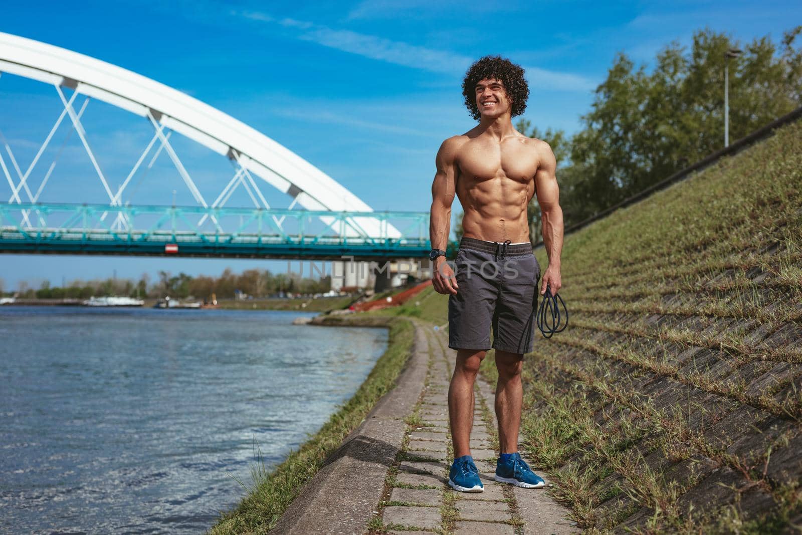 Strong bodybuilder with naked torso with six pack, perfect ABS, shoulders, biceps, triceps and chest, standing by the river, ready for morning workout. 