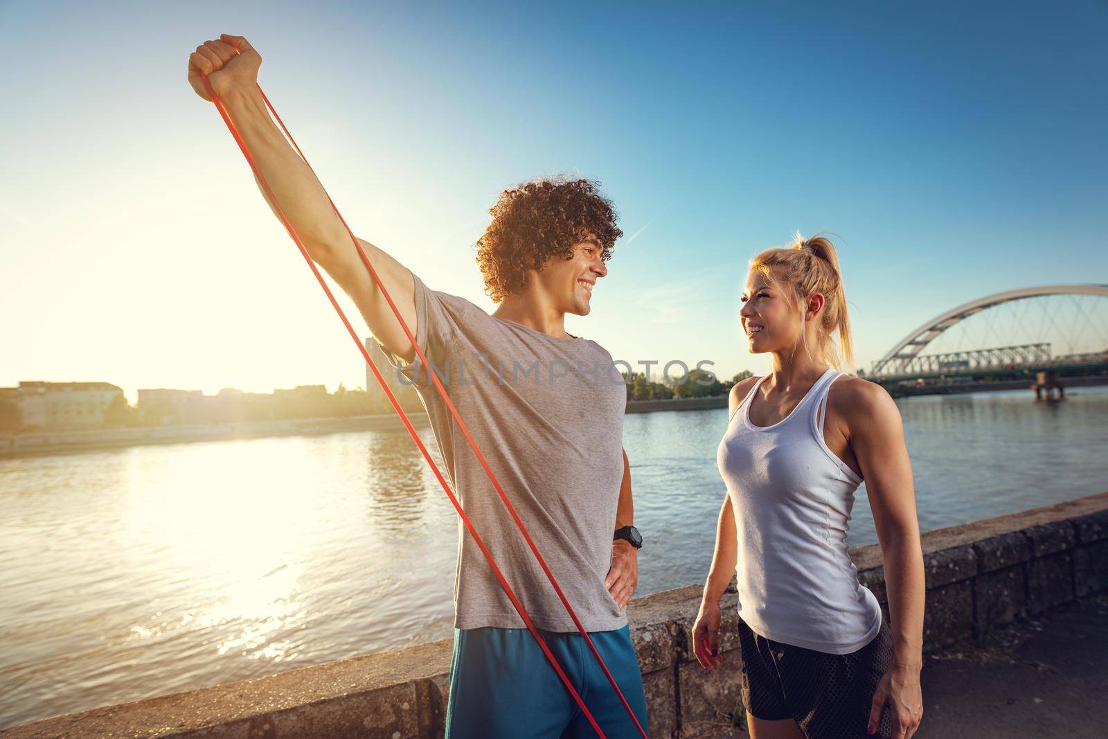 Young fitness couple doing workout with rubber band by the river in a sunset. The man is stretching arms and the woman support him.