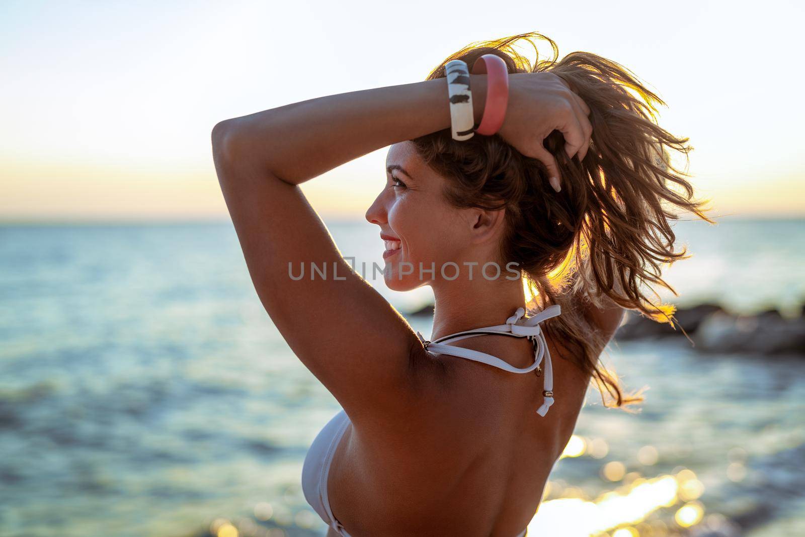 A profile of a beautiful young woman in swimsuit enjoying at sunset on the beach with raised arms. 