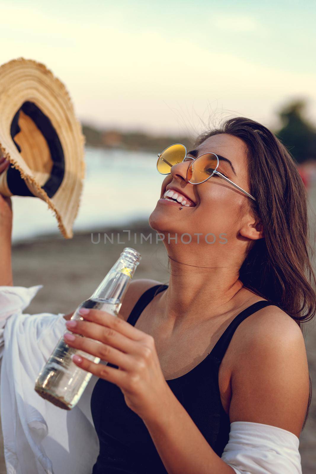 Young woman with a straw hat relaxing at sunset time on the river bank. She is sitting by the river and drinking beer. 