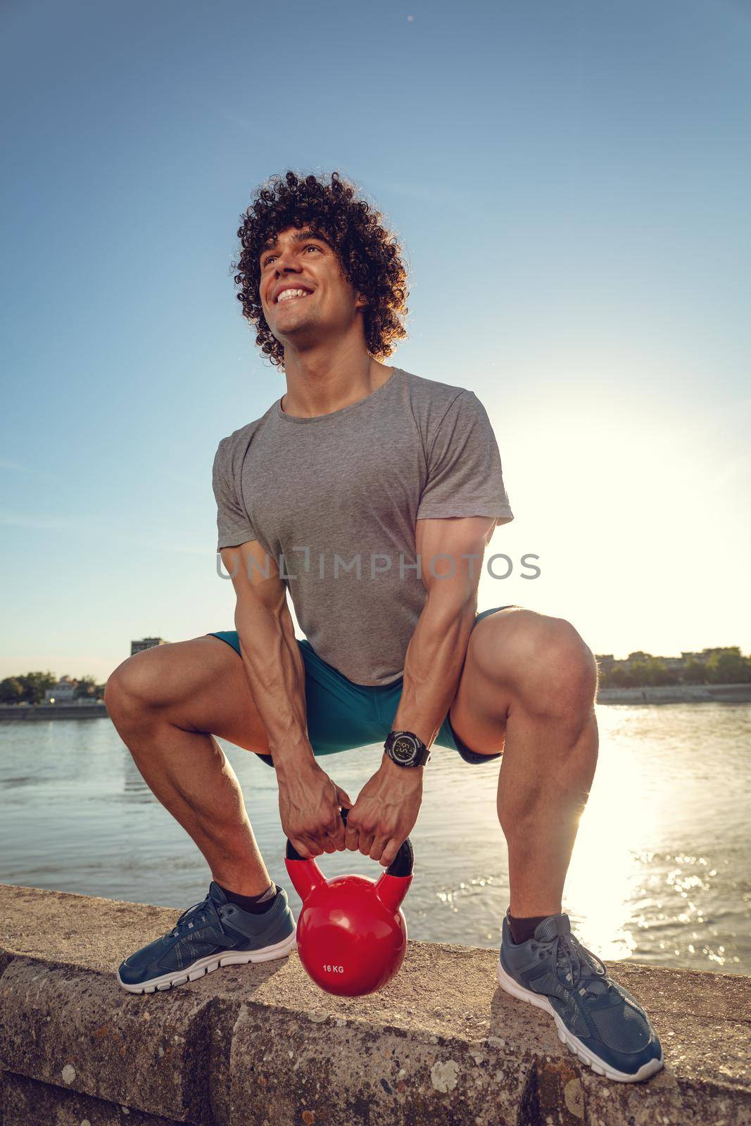 Bodybuilder is crouching and doing strong fit body training with kettlebell on the wall, near the river against blue sky with sunrise light.