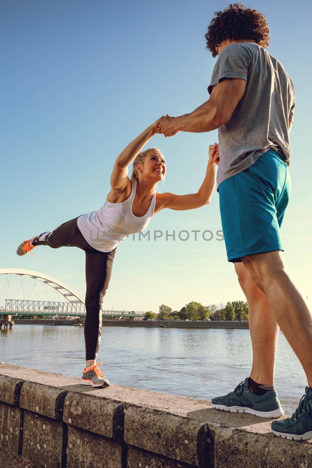 Young happy couple training outdoors by the river, stretching together on the wall at sunset.