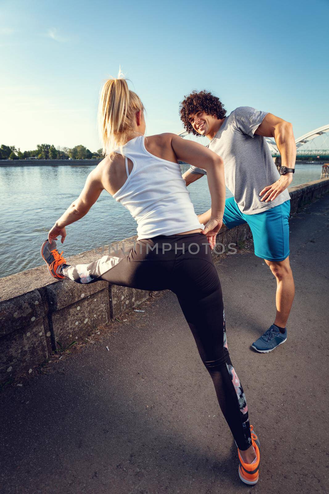 Young happy couple training outdoors by the river, stretching legs at sunset.
