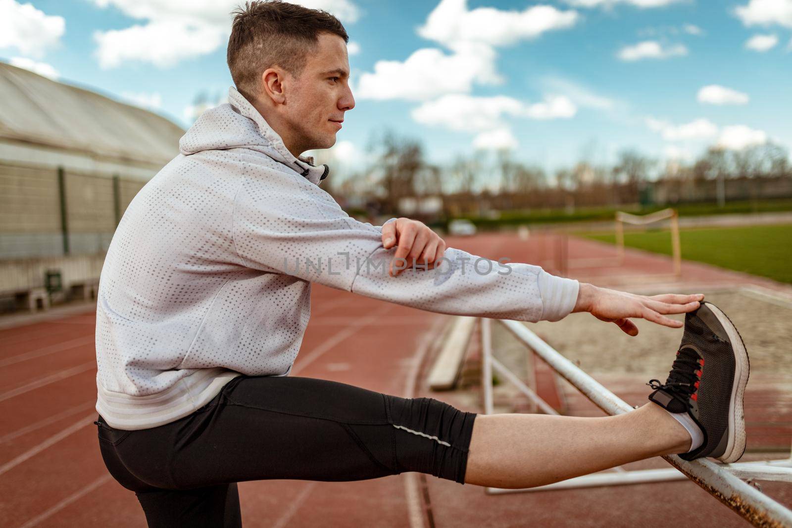 Active young man stretching and doing exercises in the public place on empty stadium. 