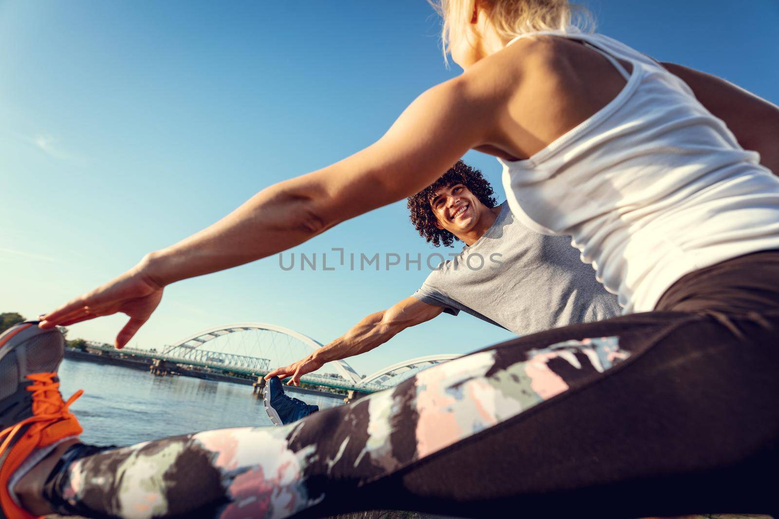 Close-up of a young happy runners couple training outdoors by the river, stretching legs at sunset.