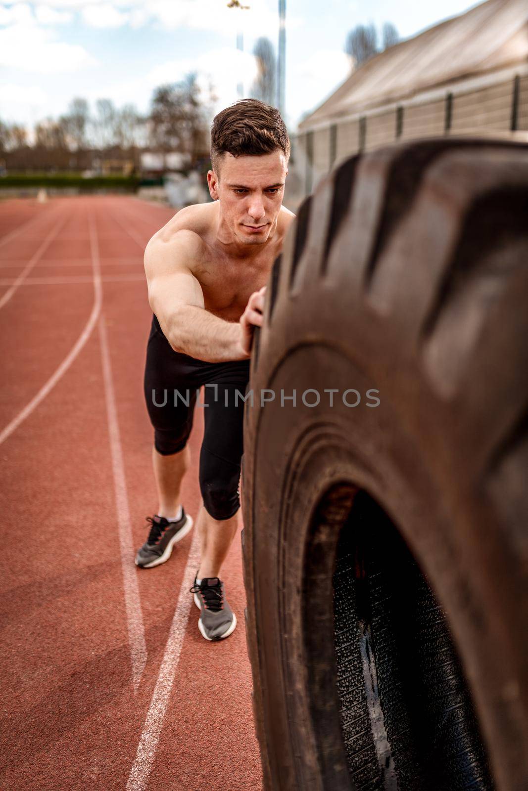 Young muscular man flipping a huge tire for training muscles at the tartan track on the stadium.