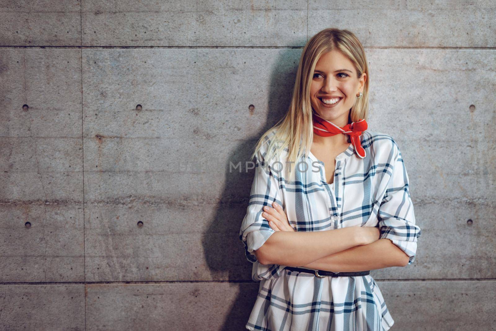 Young beautiful successful smiling female entrepreneur standing in front of a concrete wall with crossed hands and looking at camera.