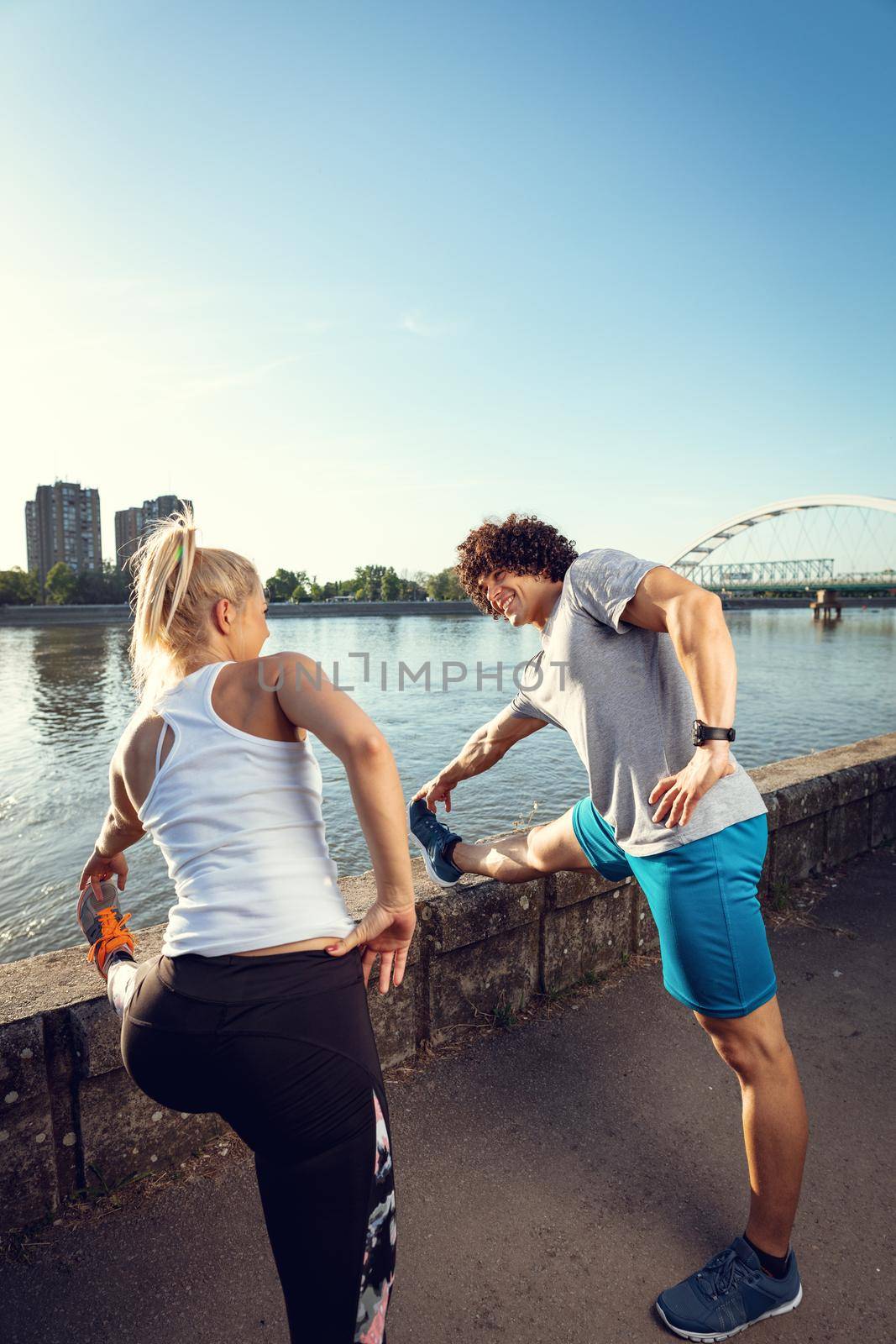 Young happy runners training outdoors by the river, stretching legs at sunset.