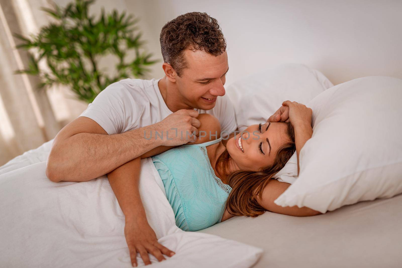 Beautiful young man waking up his cute smiling woman in bed in the morning. 