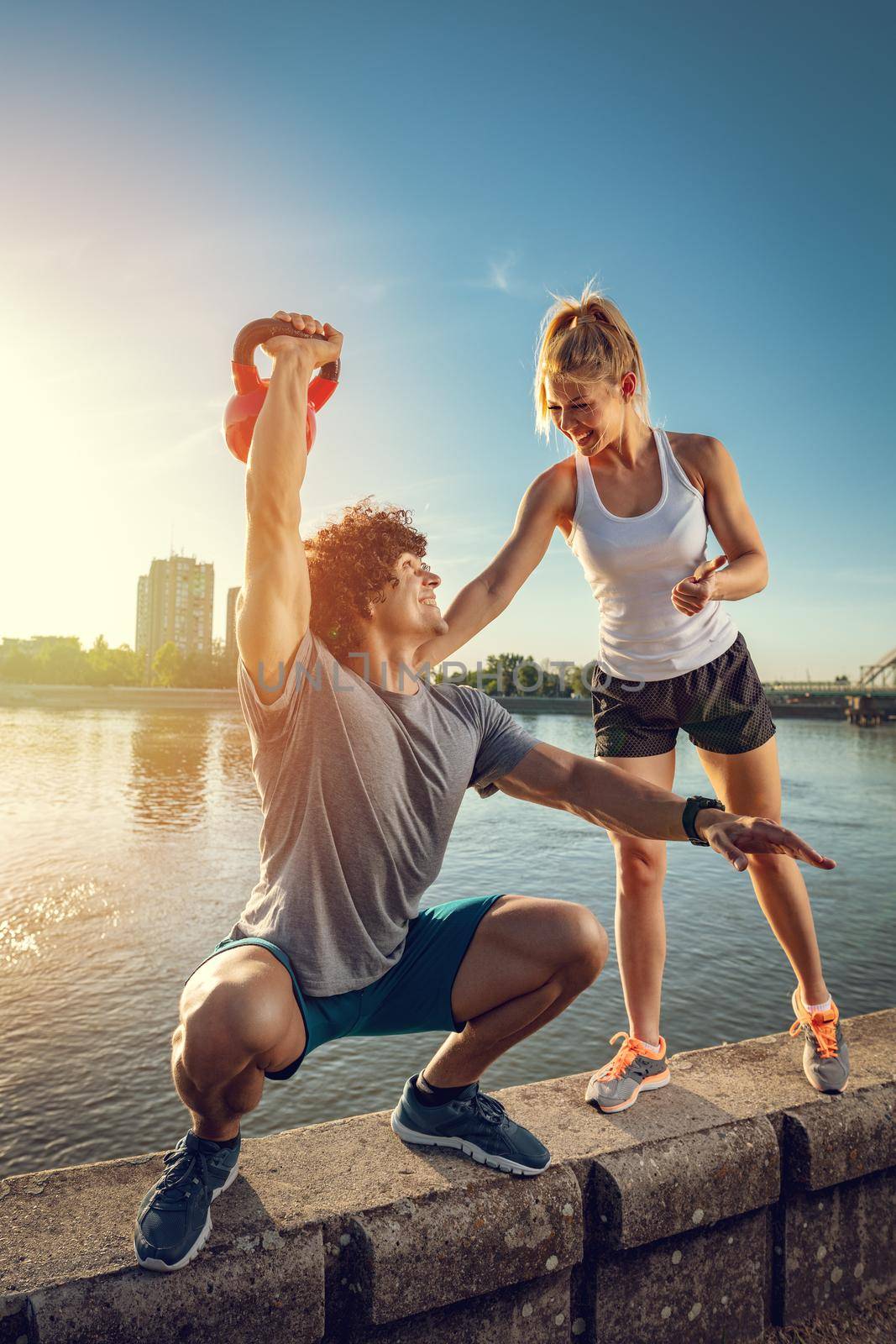 Young fitness couple doing workout with kettlebell on the wall  by the river in a sunset. The man is crouching and holding kettlebell, and the woman support him.