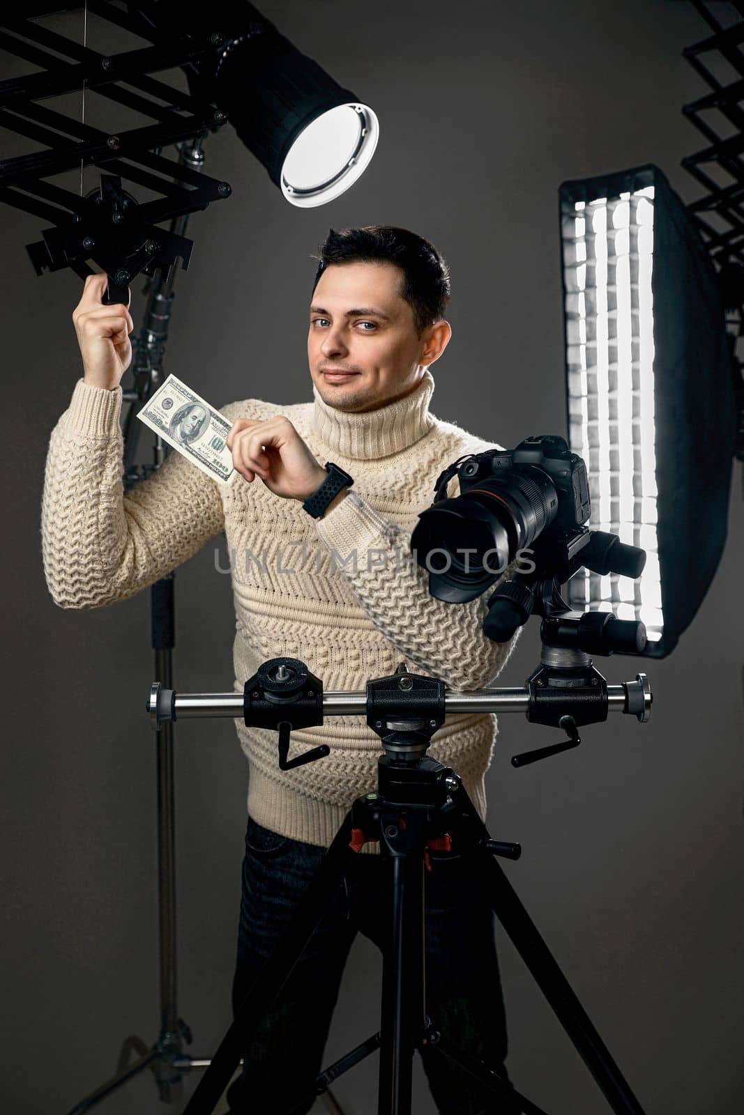 Professional caucasian photographer with digital camera on tripod holding hundred dollar bills on gray background with lighting equipment
