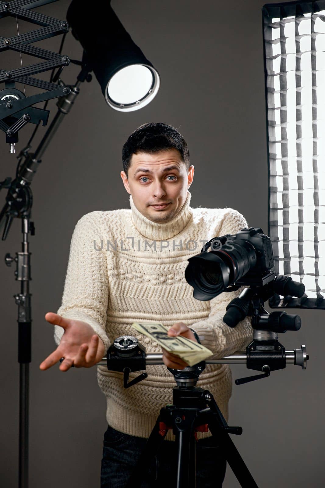 surprised photographer with digital camera on tripod holding money on gray background with lighting equipment