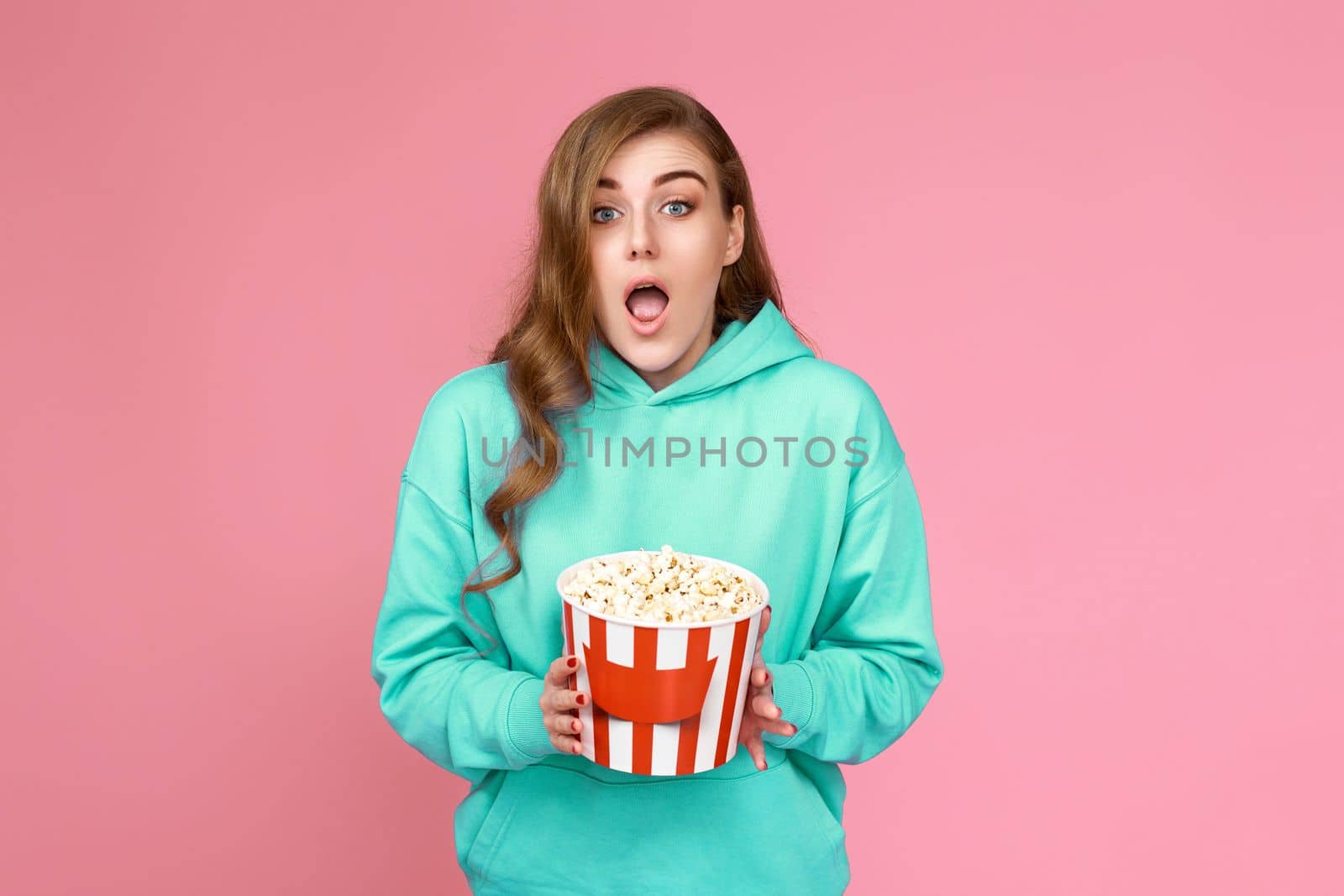amazed curly brunette woman in turquoise sweatshirt holding bucket of popcorn isolated on pink background