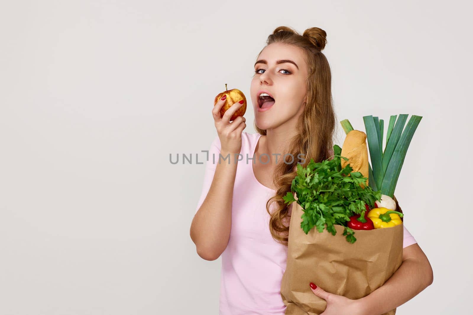 caucasian young woman in pink t-shirt with paper bag with vegetables biting apple. copy space. healthy food for diet